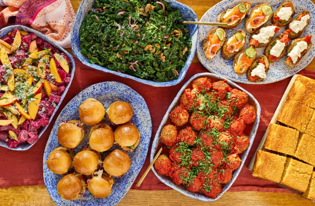 a variety of catering dishes including a roasted beat and peach salad, kale salad, fried chicken sliders, turkey meatballs and mini lox crostinis